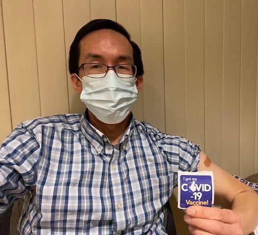 dennis-kuo a person sitting down wearing a face mask and holding a sticker that reads "I got my Covid-19 Vaccine!"