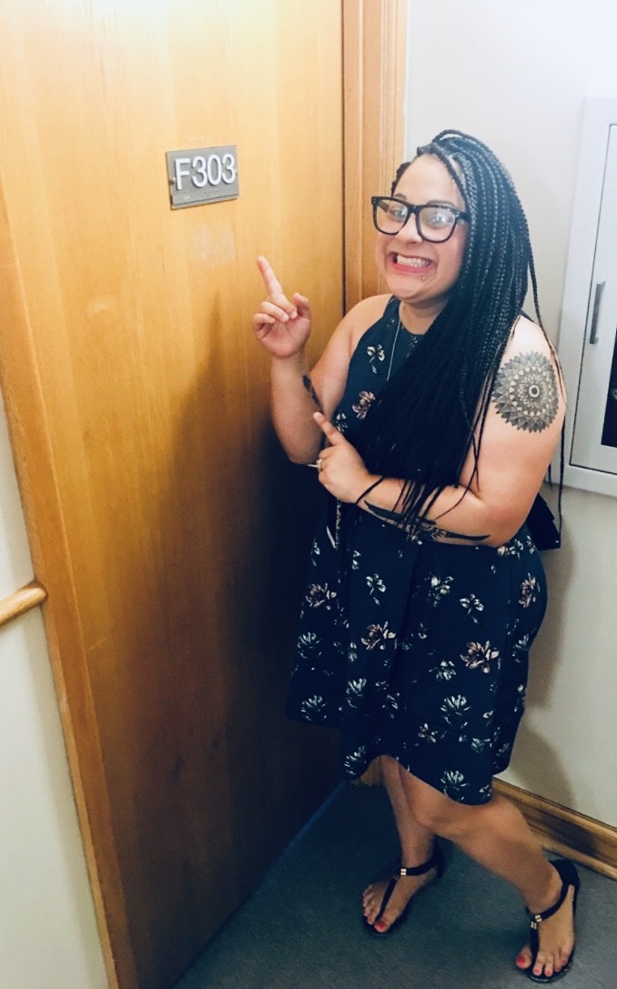 person standing by a door smiling and pointing to sign on it that reads F303