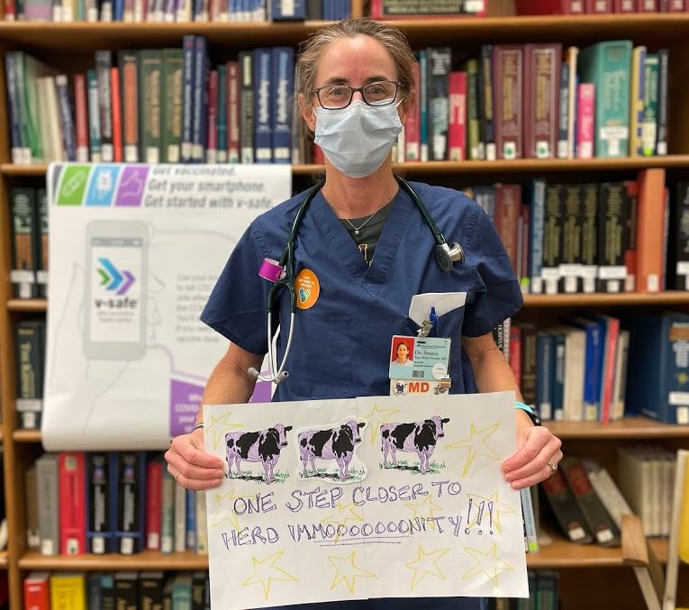 person wearing a mask and eyeglasses in a blue apron, holding a sign with cutout cows and text that reads as 'one step closer to herd immooooooonity!!!.' There is a bookshelf full of books in the background.