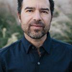 photo of a man wearing a dark blue shirt with curly hair and a beard.