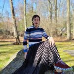 photograph of a person sitting on top of a rock. they are wearing a horizontal striped to in assorted colors, a black skirt, and maroon shoes with a multicolored edge.