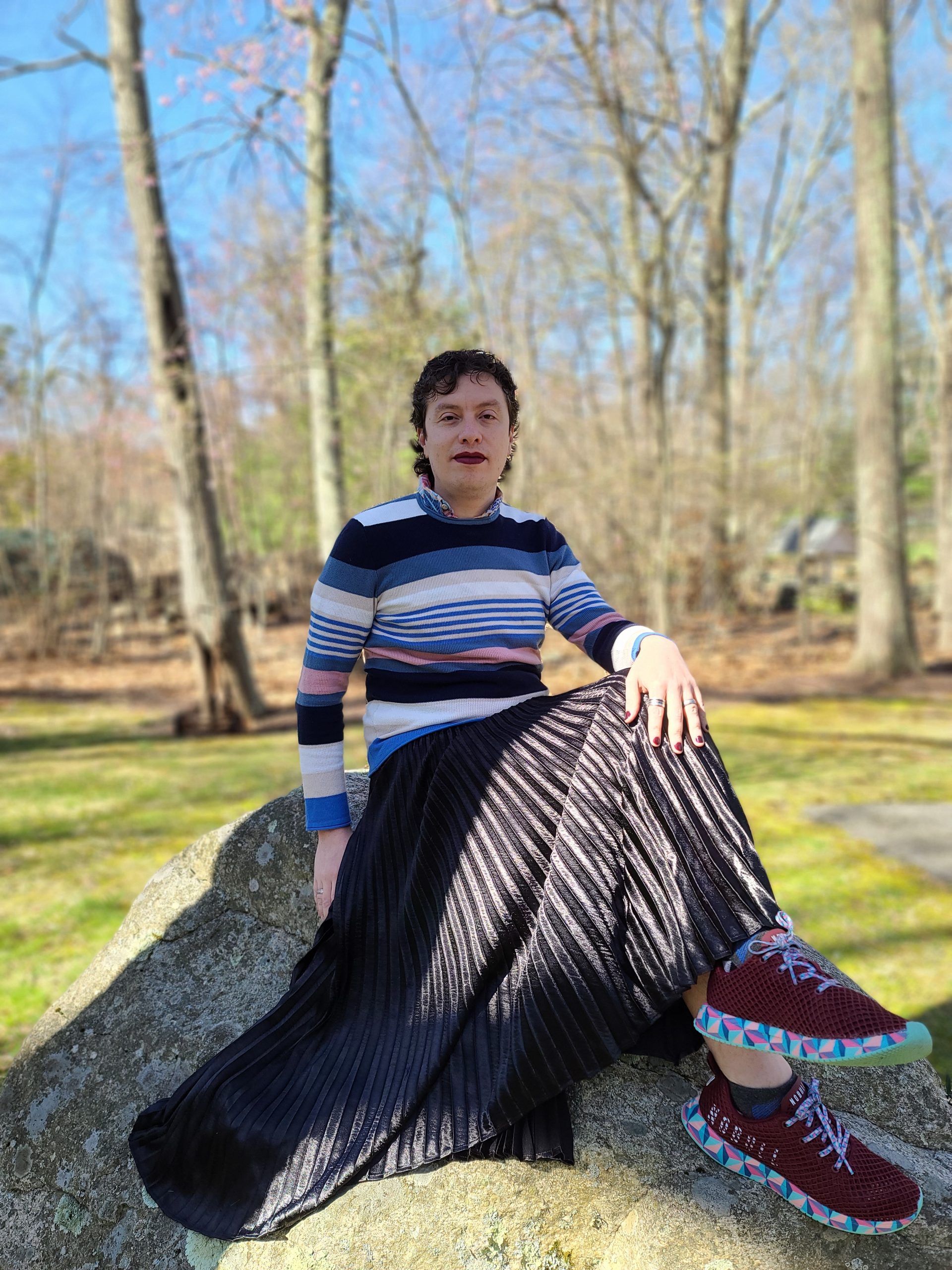 photograph of a person sitting on top of a rock. they are wearing a horizontal striped to in assorted colors, a black skirt, and maroon shoes with a multicolored edge.