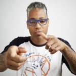 saifa flex and pointing straight ahead, wearing glasses and a shirt with words that say 'unborn, son, media.'