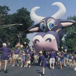 An giant purple cow balloon is carried down the street by parade volunteers wearing purple shirts.