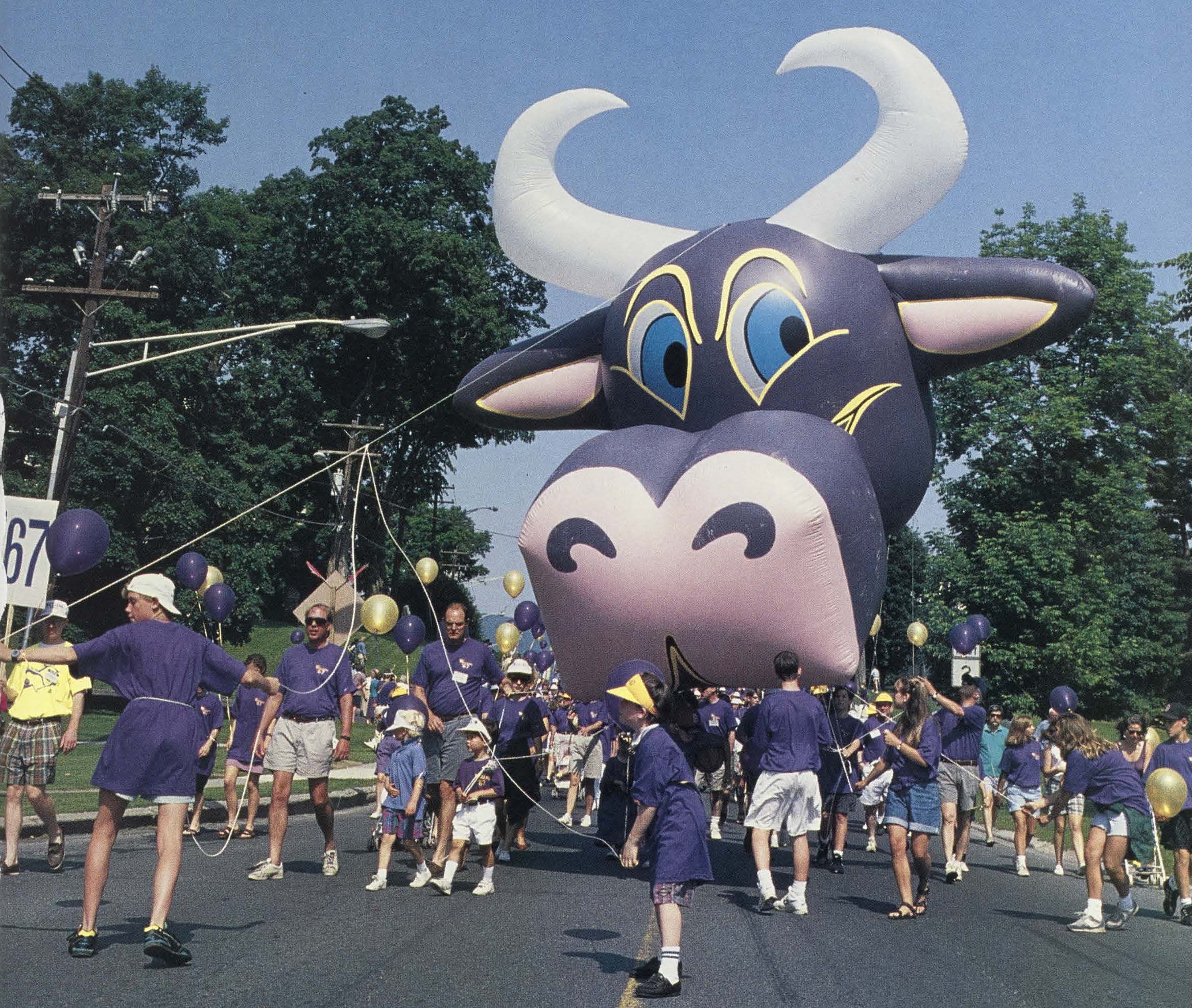 An giant purple cow balloon is carried down the street by parade volunteers wearing purple shirts.