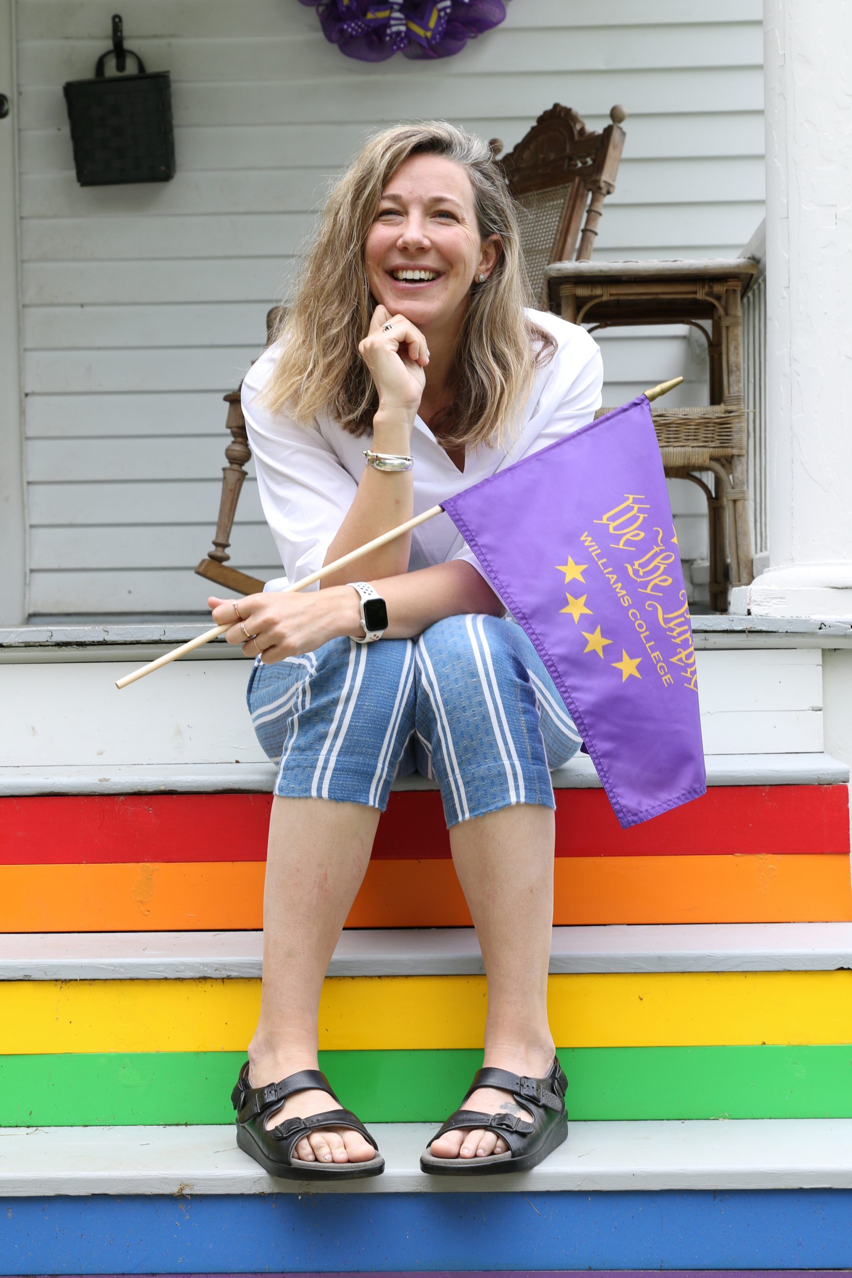 photo of a woman sitting on stairs painted with rainbow colors and holding a purple flag. She is wearing blue striped short pants, a white top, wedges, and has blond hair.