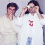 a photo of two persons playing funny poses. person on the left is wearing a white sweater and posing on a thinking pose while person on the right is wearing a white shirt that is open and wearing googly eyes
