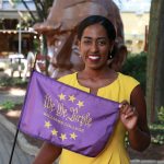 Senator Brouk poses for a photo holding a purple 'We the Purple flag in front of a head sculpture. She is wearing a yellow dress.