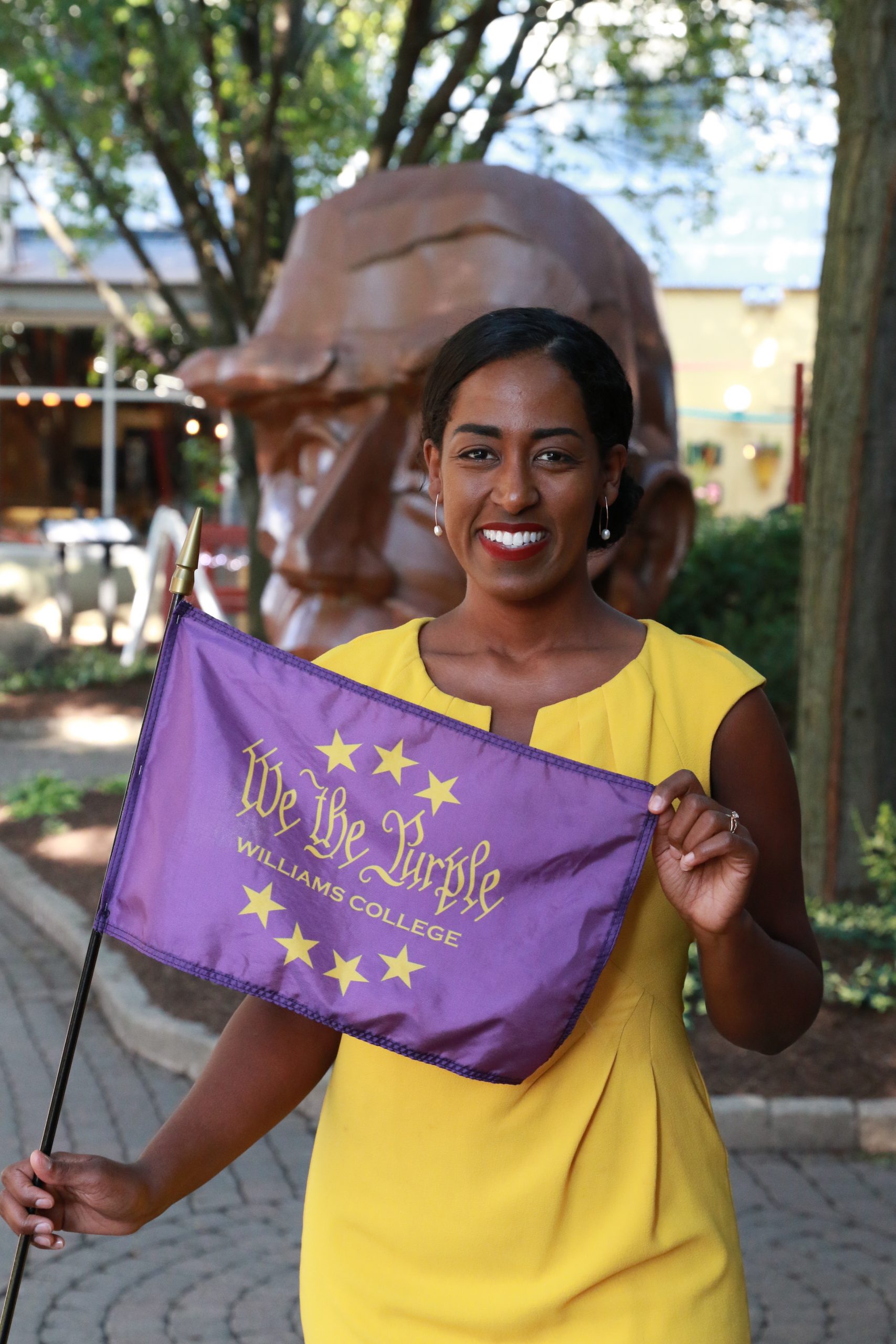 Senator Brouk poses for a photo holding a purple 'We the Purple flag in front of a head sculpture. She is wearing a yellow dress.