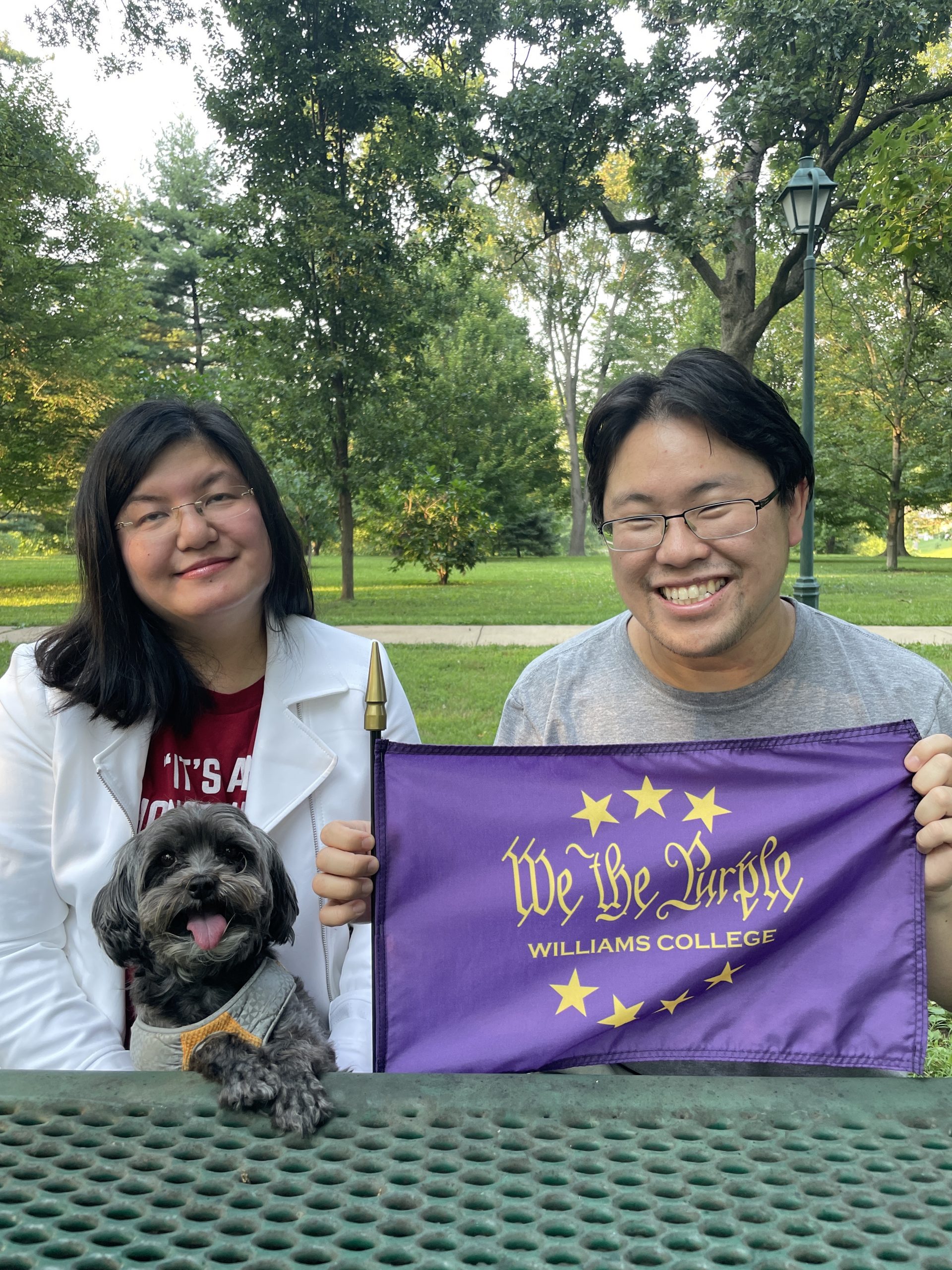 Caroline, Kevin, and Pixie holding a We the Purple flag while sitting down at a park table.