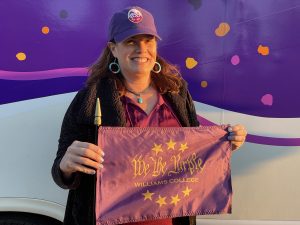 a woman holding a purple falg that says 'We the Purple.' She is wearing a maroon top with a purple hat and black coat.