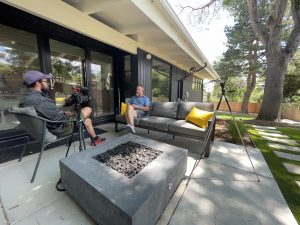 2 people sitting outside on a concrete deck with a fireplace in the center and house portion on the left.
