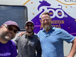 three people posting for a group photo side by side in front of an RV with a purple cow silhouette decal that reads "Williams Alumni."