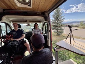 a view inside a van of three people conducting an on-camera interview with the right half showing a landcape view with blue and cloudy sky and a tree