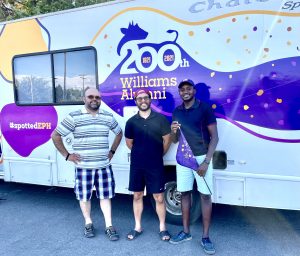 3 people standing for group photo in front of an RV decorated with a purple cow logo with text that says 200th Williams Alumni
