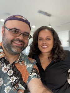2 people posing for a selfie. Person on the left is a man wearing a baseball cap backwards, glasses, and flower patterned shirt. Person on the right is a woman with long hair, wearing a balck top.