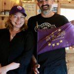 Aaron and his wife, Jasmine, posing for a photo inside Old Home Distillers' tasting room. Jasmine is standing to the left of Aaron and is wearing a black top with a purple Williams bicentennial baseball cap, with her hands crosses. Aaron is standing to Jasmine's right while holding a 'We the Purple' purple flag and wearing a black Old Home Distillers branded shirt and a Williams bicentennial baseball cap.