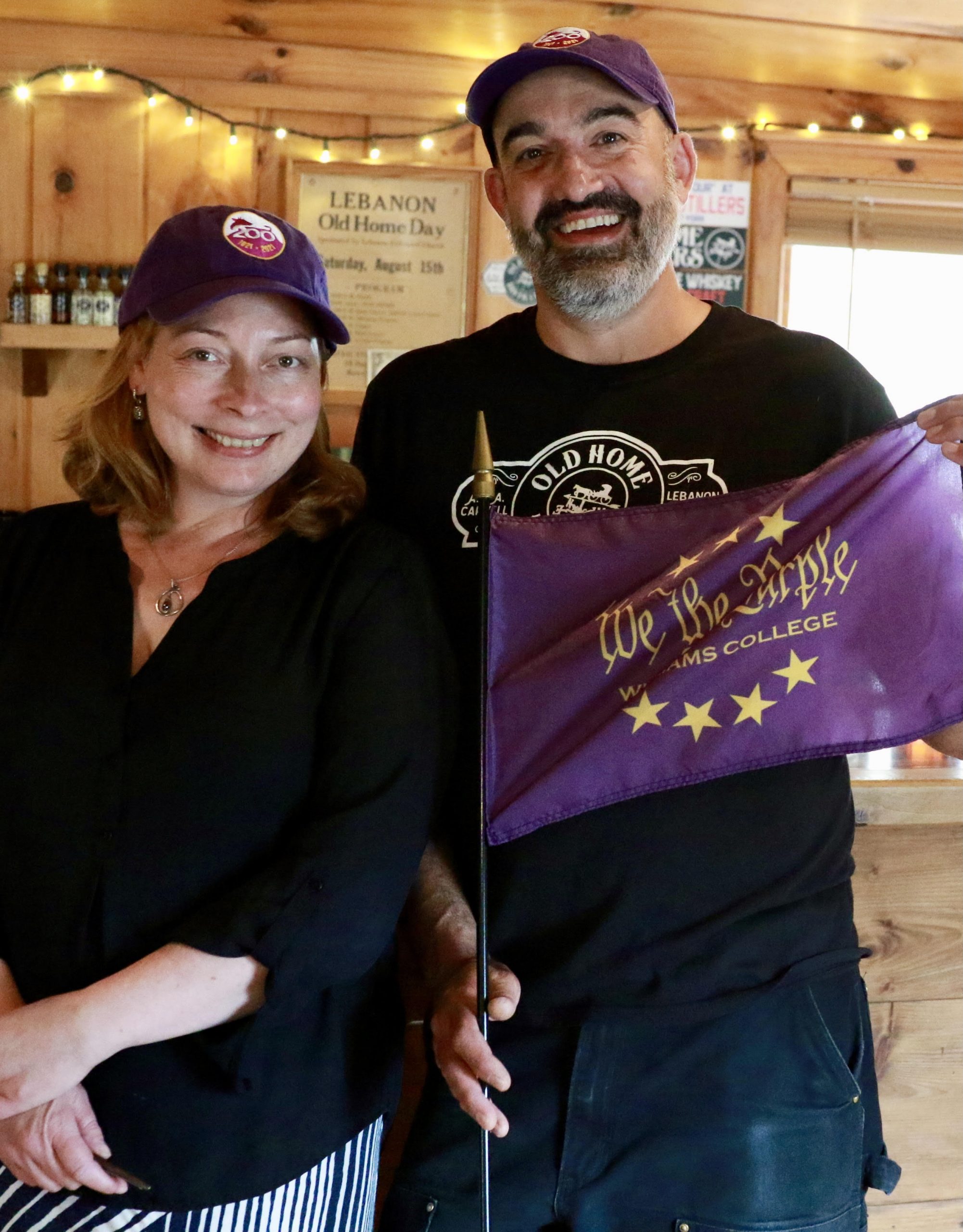 Aaron and his wife, Jasmine, posing for a photo inside Old Home Distillers' tasting room. Jasmine is standing to the left of Aaron and is wearing a black top with a purple Williams bicentennial baseball cap, with her hands crosses. Aaron is standing to Jasmine's right while holding a 'We the Purple' purple flag and wearing a black Old Home Distillers branded shirt and a Williams bicentennial baseball cap.