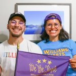 A portrait photograph of Kevin and Alejandra sitting down on a couch, holding a purple 'We the Purple' flag, with a frame of a landscape photo of a lake in the background. Kevin is on the left wearing a black baseball cap with a logo of the bicentennial cow 200th and is wearing a white shirt that has Williams in pride colors. Alejandra is sitting to the right wearing a purple baseball cap backwards and is wearing a blue shirt that reads with yellow text as Williams Firsts.
