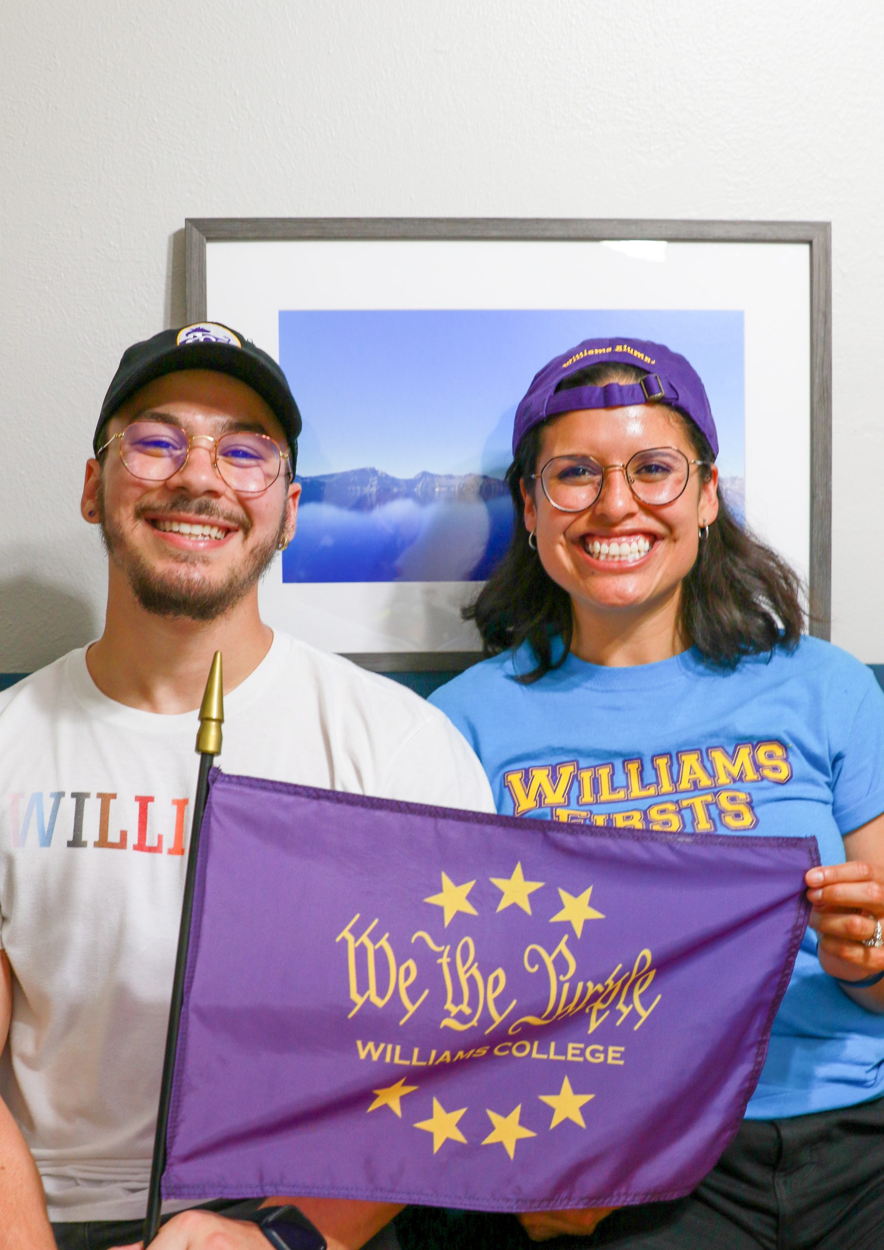 A portrait photograph of Kevin and Alejandra sitting down on a couch, holding a purple 'We the Purple' flag, with a frame of a landscape photo of a lake in the background. Kevin is on the left wearing a black baseball cap with a logo of the bicentennial cow 200th and is wearing a white shirt that has Williams in pride colors. Alejandra is sitting to the right wearing a purple baseball cap backwards and is wearing a blue shirt that reads with yellow text as Williams Firsts.