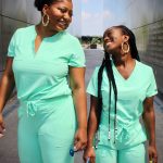 two Black doctors in their green scrubs posing standing side by side and smiling at each other.