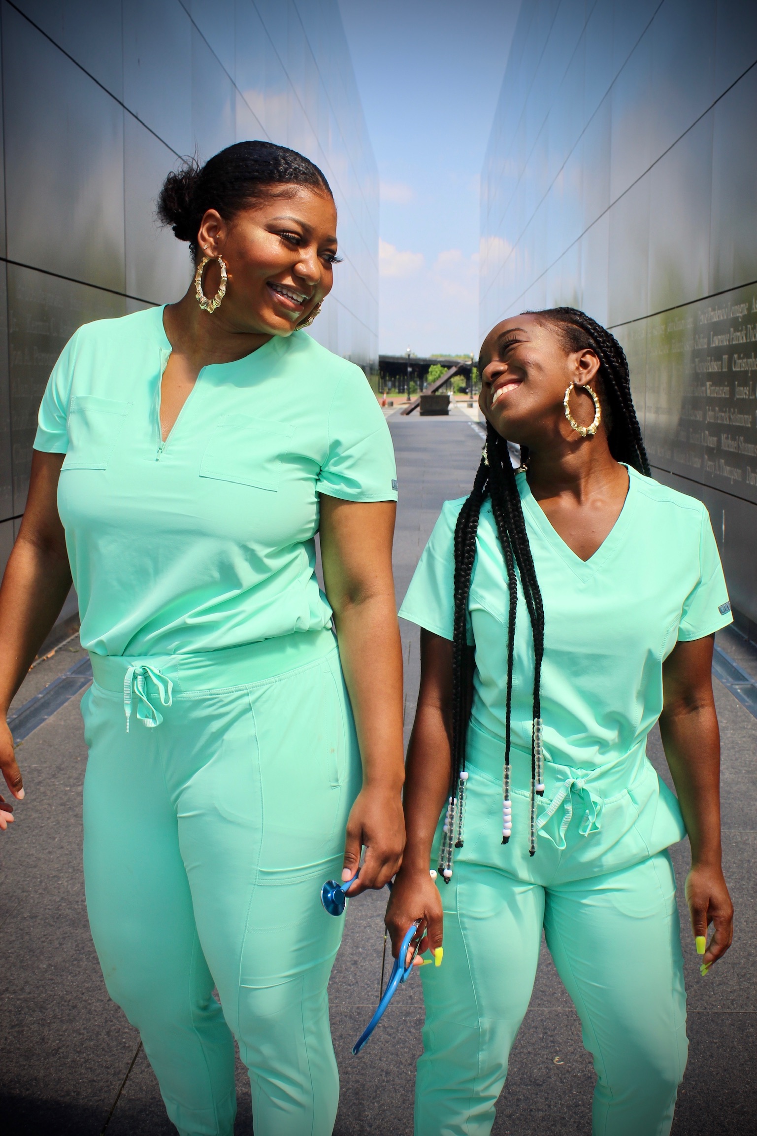 two Black doctors in their green scrubs posing standing side by side and smiling at each other.
