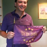 a man, standing up wearing a purple polo shirt and white shorts while holding a purple flag that says 'We the Purple'