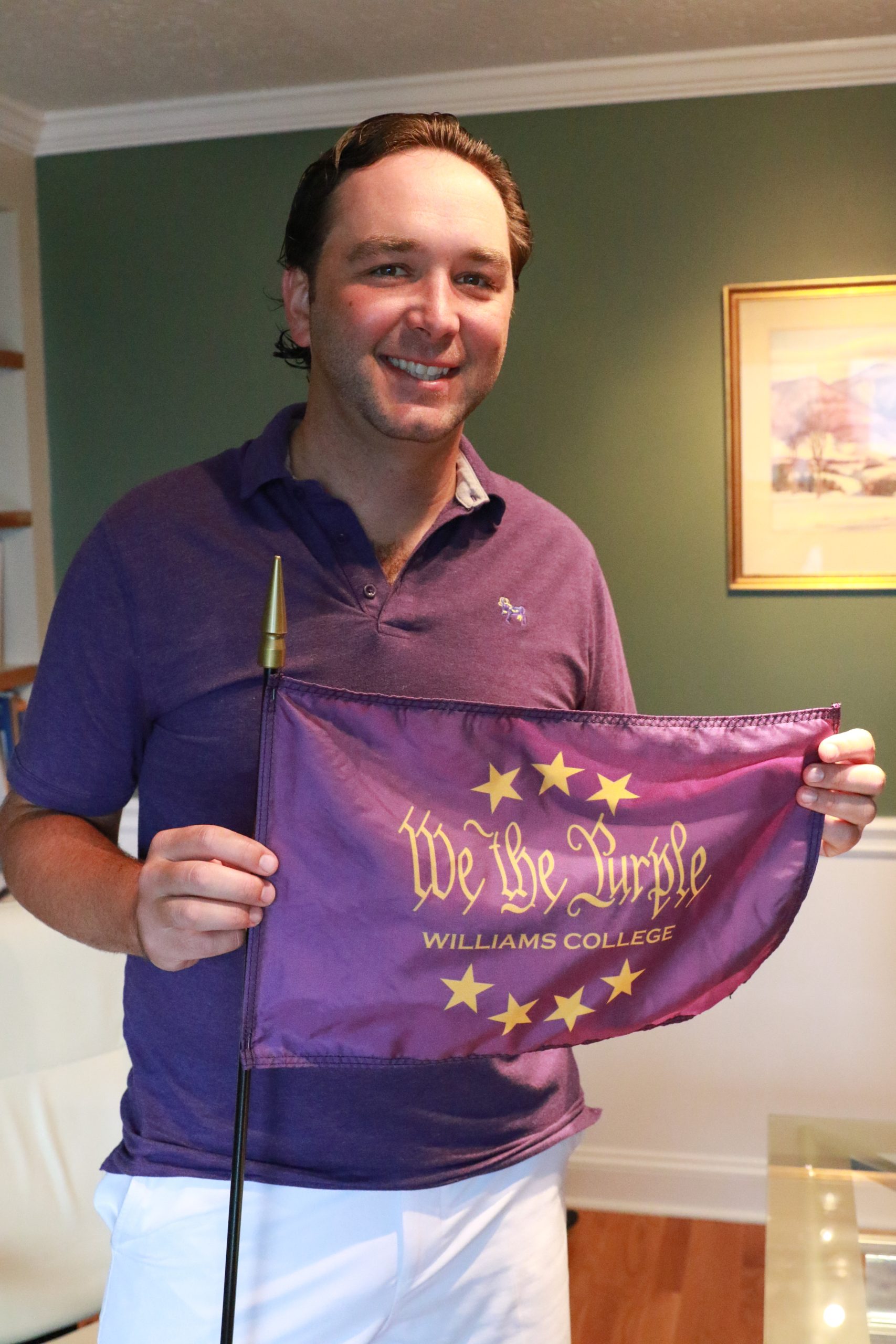 a man, standing up wearing a purple polo shirt and white shorts while holding a purple flag that says 'We the Purple'