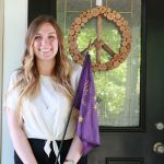 Alex holding 'We the Purple' flag while standing in front of a door with a peace sign wood decoration
