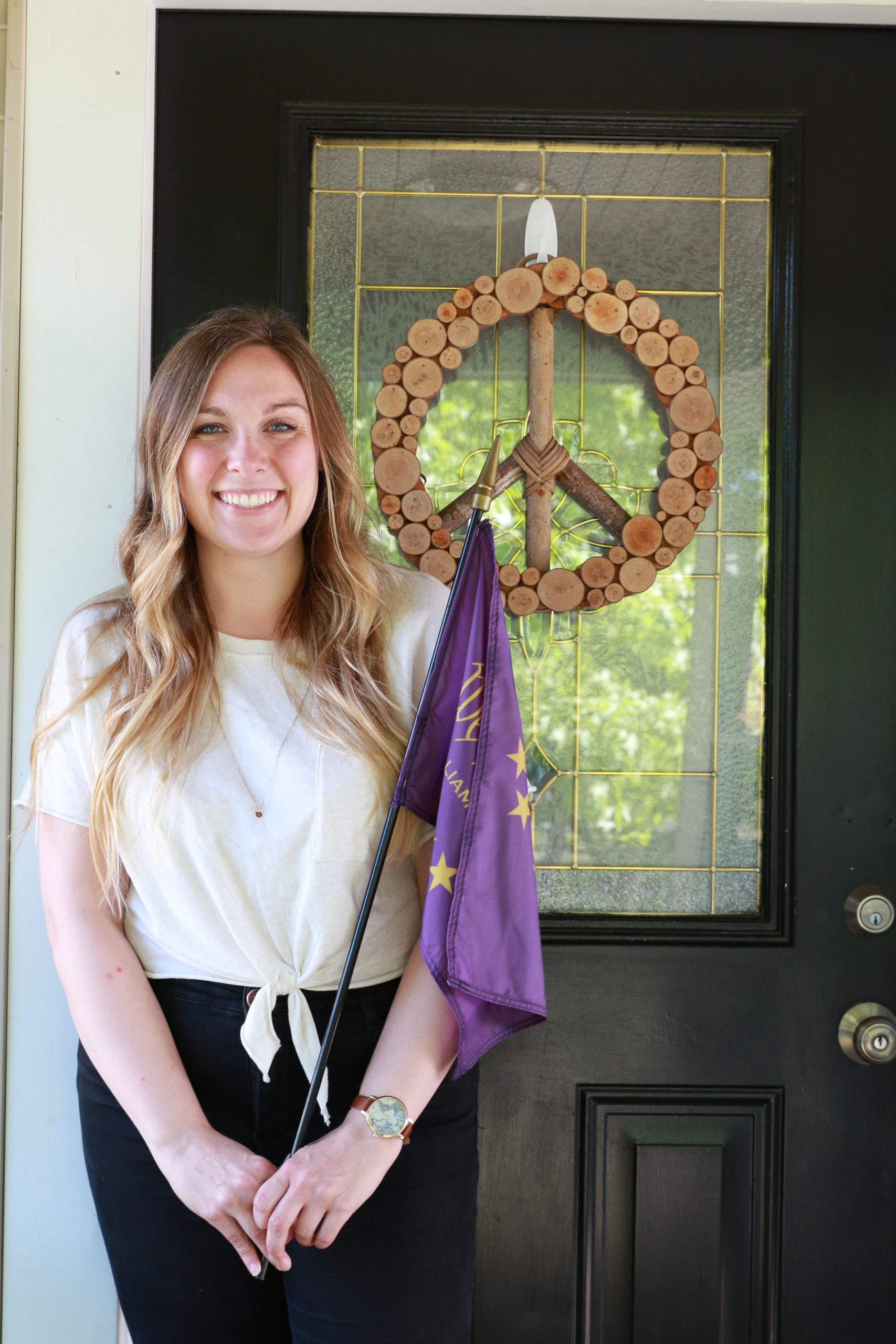 Alex holding 'We the Purple' flag while standing in front of a door with a peace sign wood decoration