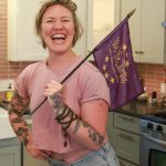 a woman standing on a kitchen holding a purple flag that has text reading 'We the Purple.' She is wearing a light pink top, blue jeans, and has tattoos on her arms.