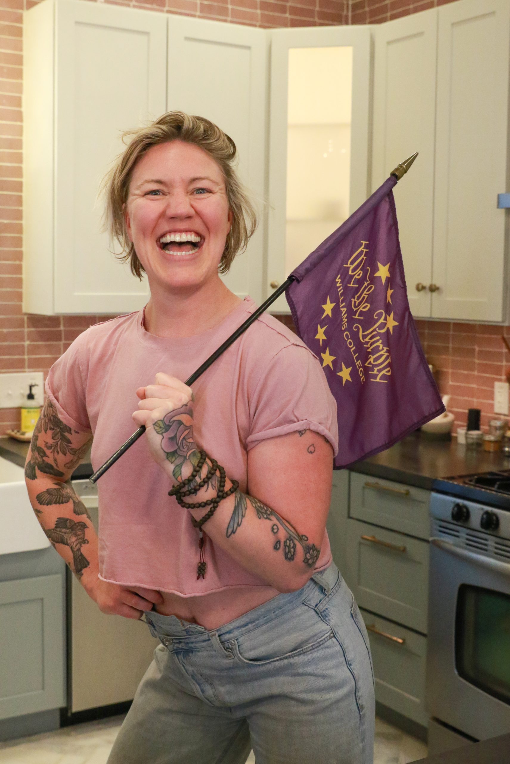 a woman standing on a kitchen holding a purple flag that has text reading 'We the Purple.' She is wearing a light pink top, blue jeans, and has tattoos on her arms.
