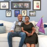 Tamaan and Camille sitting down on their living room sofa with a backdrop of their family portraits on the wall. Camille is holding a 'We the Purple' flag.