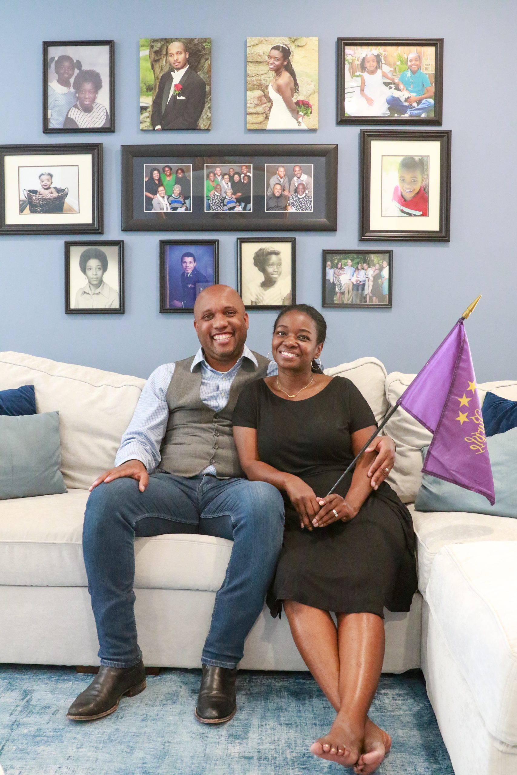 Tamaan and Camille sitting down on their living room sofa with a backdrop of their family portraits on the wall. Camille is holding a 'We the Purple' flag.