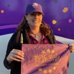 a woman standing and holding a purple flag that says 'We the Purple' She is wearing a purple hat, maroon top, and black coat with a purple hat.