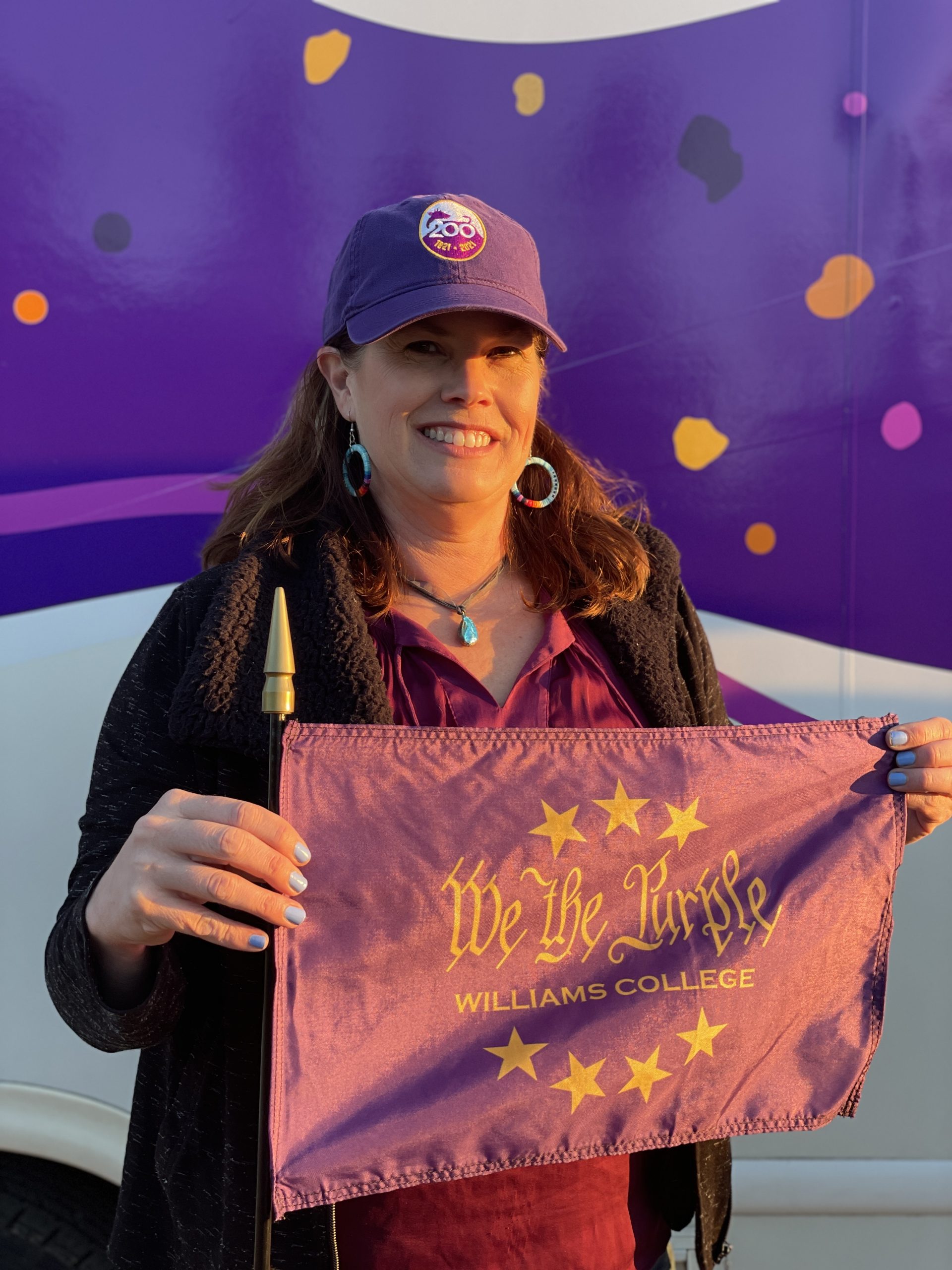 a woman standing and holding a purple flag that says 'We the Purple' She is wearing a purple hat, maroon top, and black coat with a purple hat.