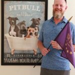 person standing next to a poster depicting a group of 4 pitbulls inside a bathtub with text that reads ' Pitbull and Co Bath Soap - Wash your Paw'
