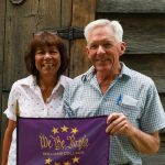 2 people standing in front of a weathered barn door while holding a purple flag that says We the Purple