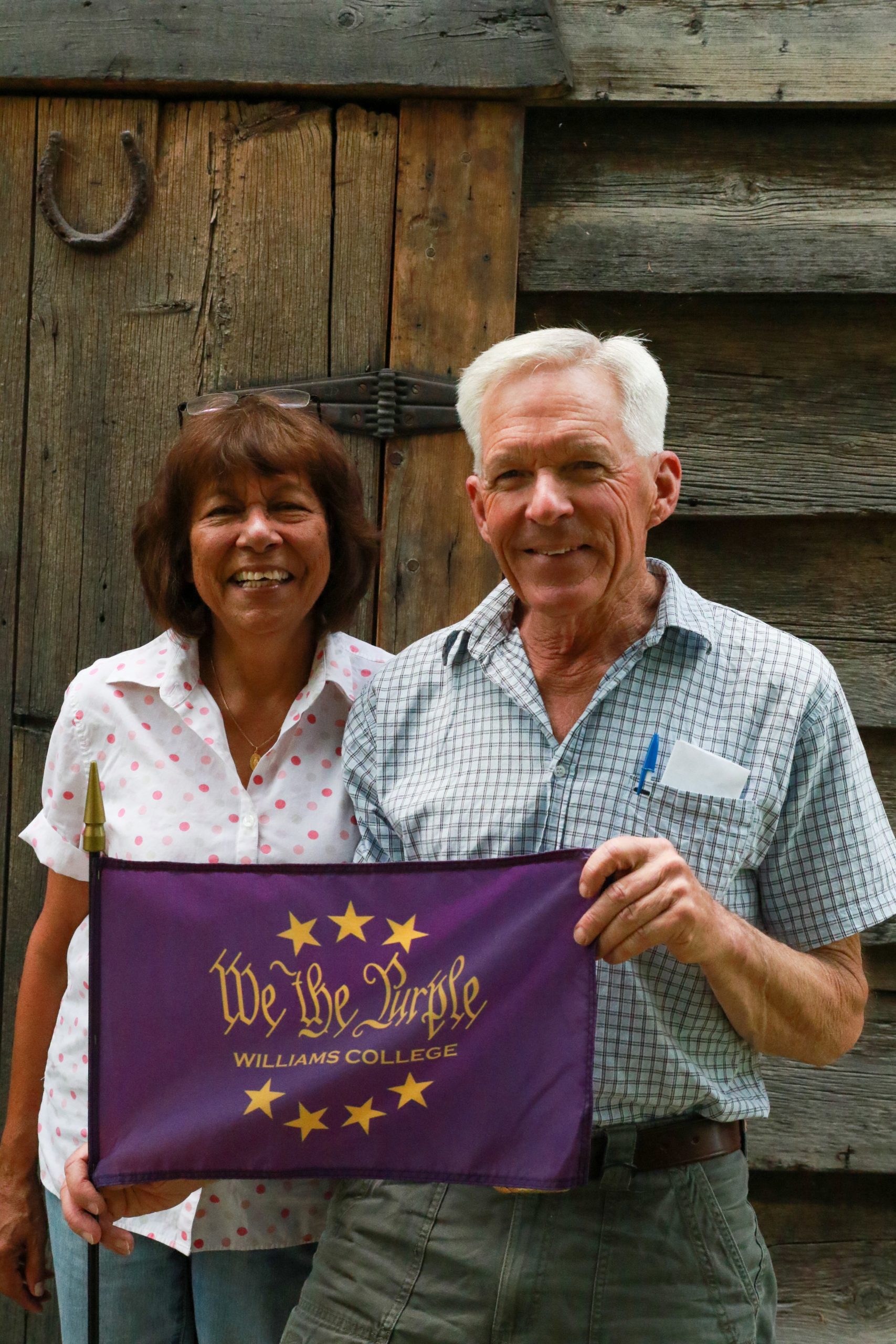 2 people standing in front of a weathered barn door while holding a purple flag that says We the Purple