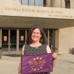 a person standing outside a museum entrance holding a purple flag that says 'We the Purple' and lettering above the entrance reads 'Natural History Museum of Utah'