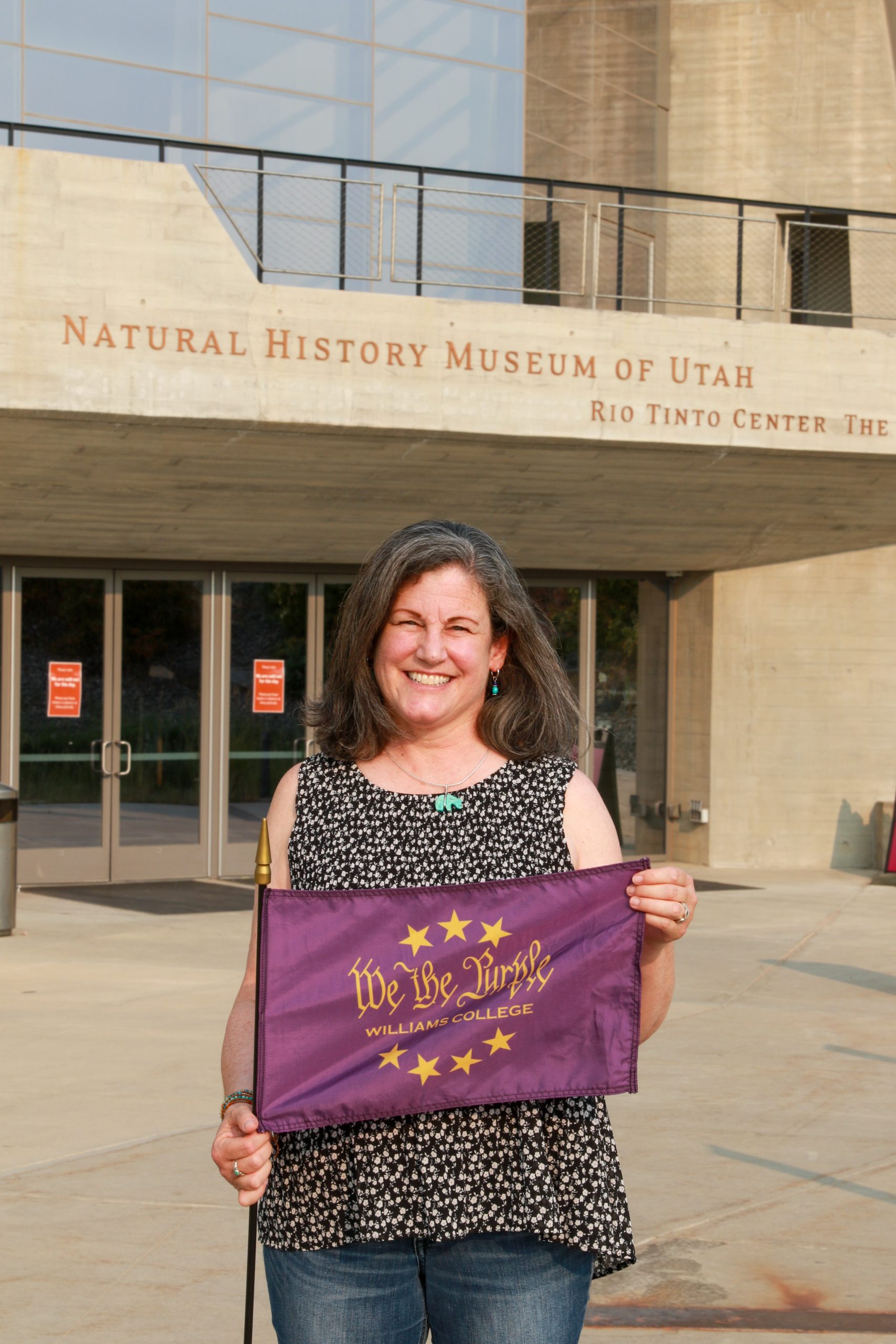 a person standing outside a museum entrance holding a purple flag that says 'We the Purple' and lettering above the entrance reads 'Natural History Museum of Utah'