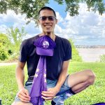 man kneeling by a purple picnic blanket in a park while holding a purple flag with a purple hat at the top