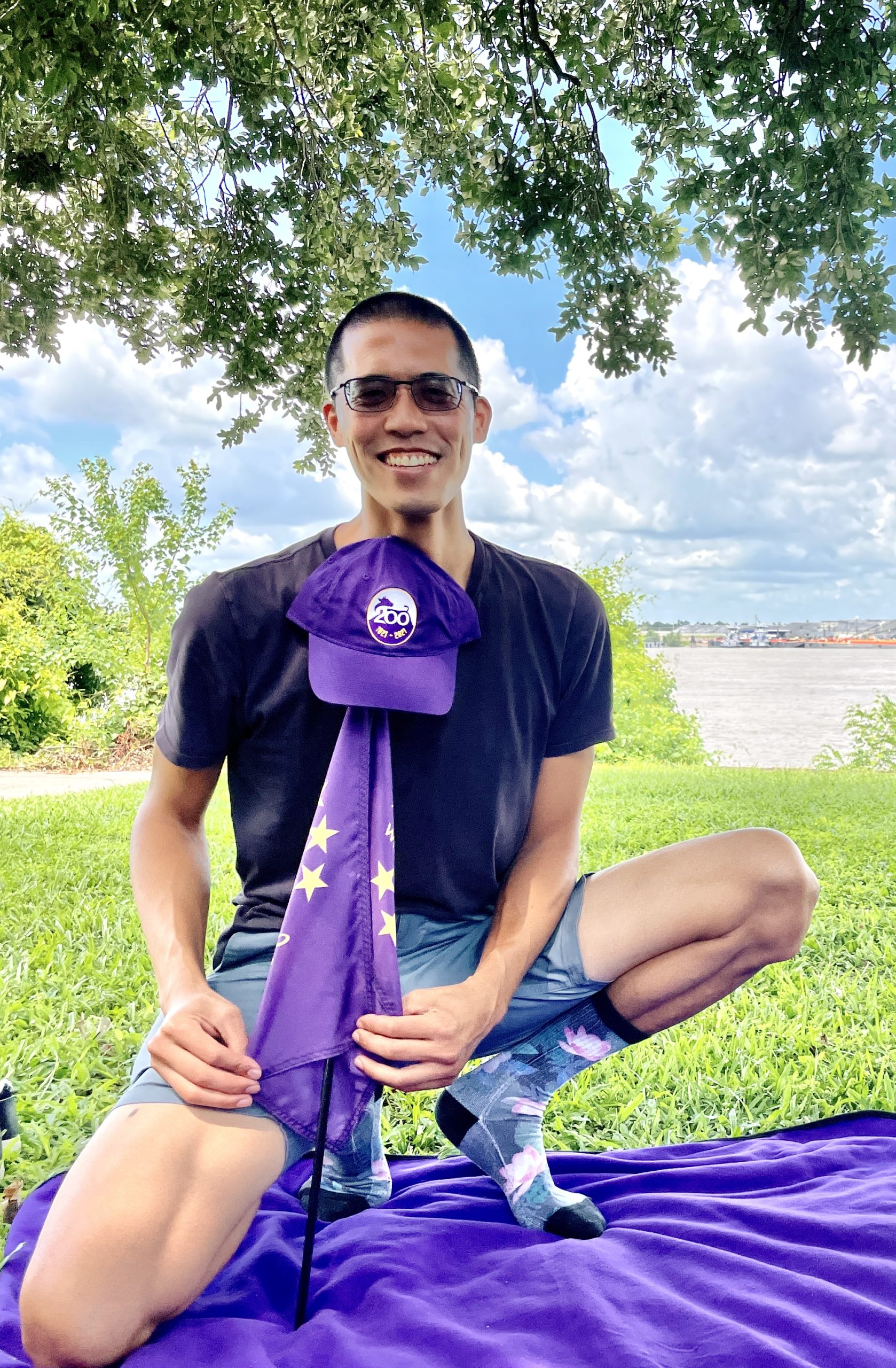 man kneeling by a purple picnic blanket in a park while holding a purple flag with a purple hat at the top