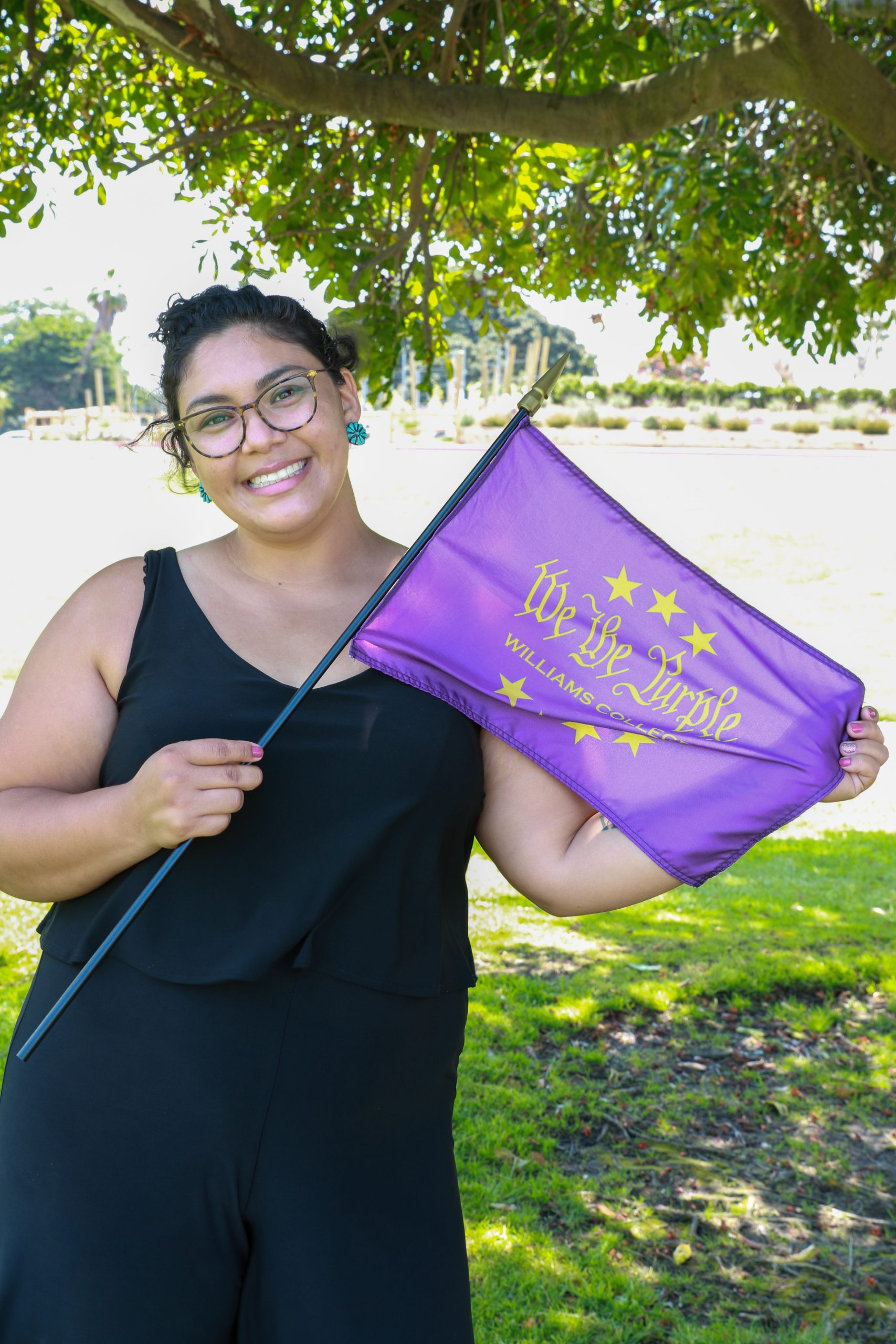 a woman wearing a baclk dress posing for a photo in a park while holding a purple flag that says 'We the Purple'