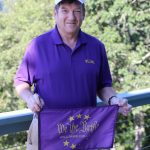 a man dressed in tan shorts and a purple shirt, with a white baseball cap with letters that read 'rugby' holding a purple flag that says 'We the Purple