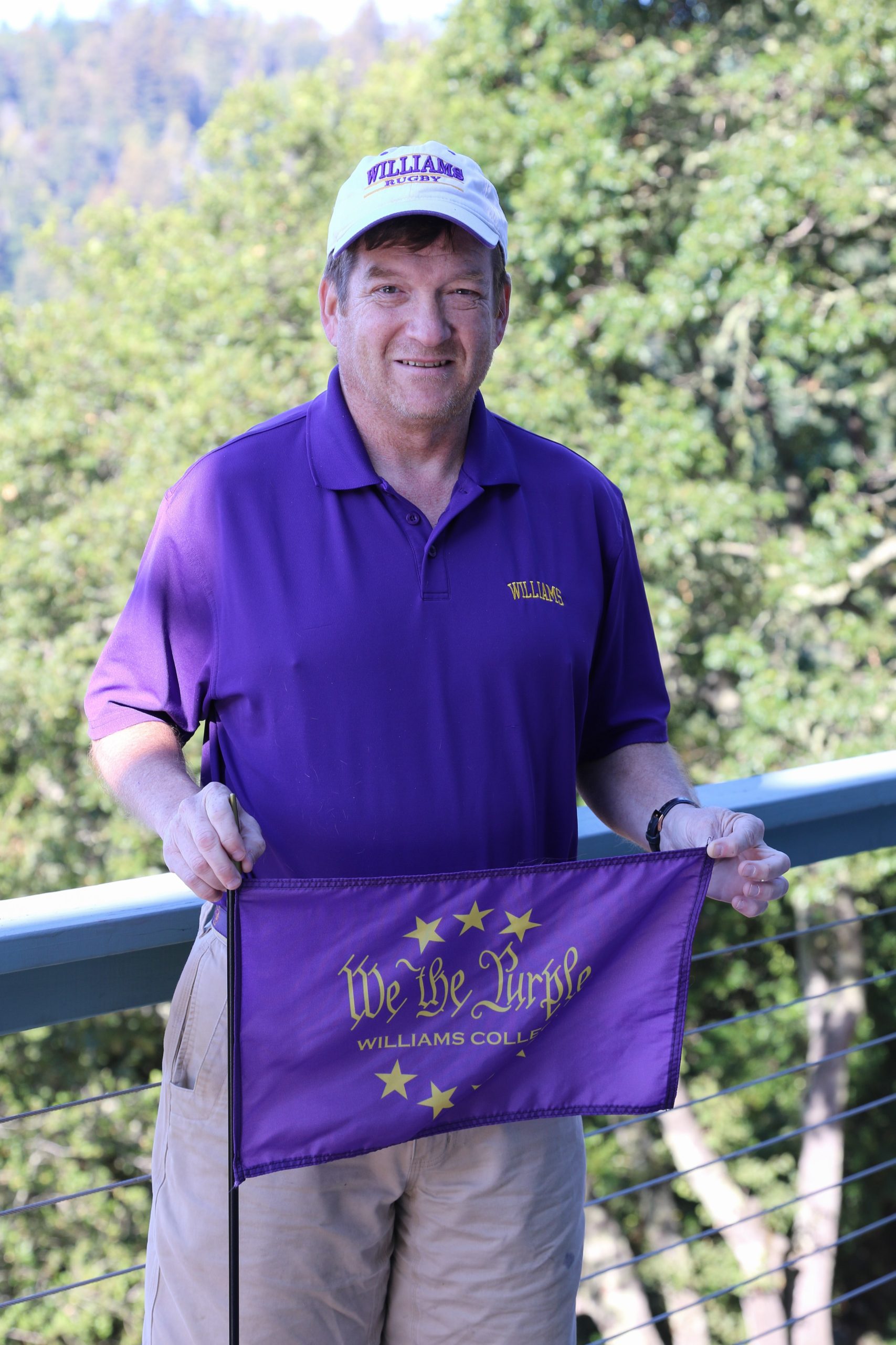 a man dressed in tan shorts and a purple shirt, with a white baseball cap with letters that read 'rugby' holding a purple flag that says 'We the Purple