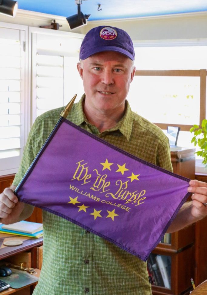 a man wearing a green shirt and holding a purple flag that says 'We the Purple, Williams College," and is also wearing a purple hat with a cow logo.