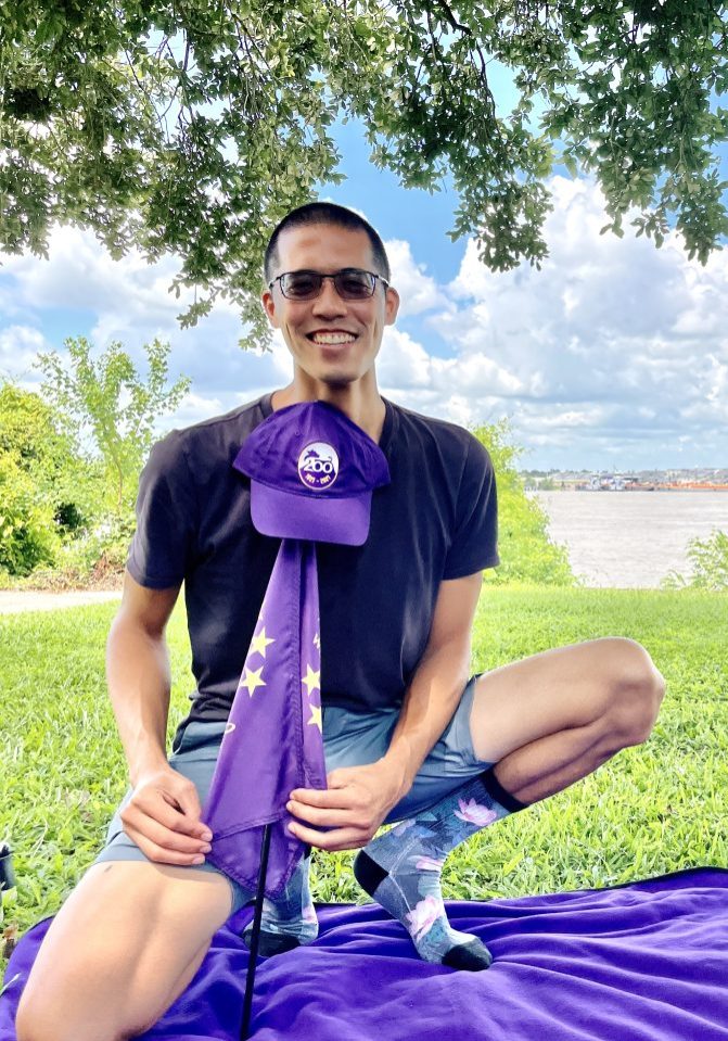 man kneeling by a purple picnic blanket in a park while holding a purple flag with a purple hat at the top