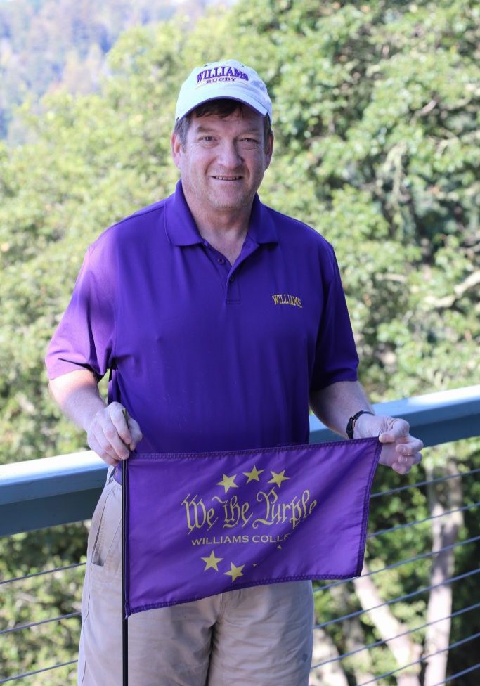 a man dressed in tan shorts and a purple shirt, with a white baseball cap with letters that read 'rugby' holding a purple flag that says 'We the Purple