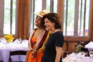 Edwina Martin and Williams College President Maud S. Mandel stand close together, smiling as they pose for a photo in a warmly lit dining hall. Edwina wears a bright orange outfit with a stylish hat and a colorful scarf, while President Mandel wears a navy blue dress with a silver necklace. Tables in the background are set with white tablecloths, folded napkins, and glassware, with vases of yellow flowers adding a cheerful touch. Large windows allow natural light to filter into the room. 